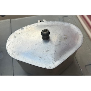May include: A silver-colored metal mess kit lid with a weathered, textured surface and a small, black, cylindrical knob centered on top. The lid has an irregular, rounded triangular shape with a small metal tab extending from one edge.
