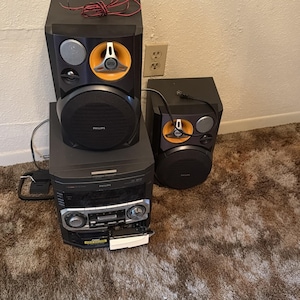 May include: A black Philips stereo system featuring a central unit with a cassette deck and two speakers. Each speaker has a large black woofer, a silver tweeter, and a prominent yellow circular accent around the upper driver. Wires are attached.