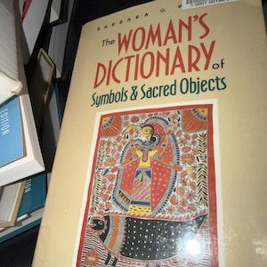 May include: A beige paperback book titled The Woman's Dictionary of Symbols & Sacred Objects by Barbara G. Walker. The cover features a colorful, intricate folk-art illustration of a central figure surrounded by floral patterns and a black animal.