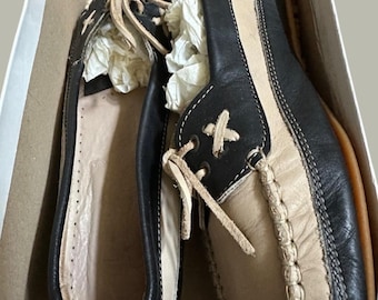 May include: A pair of two-tone moccasins featuring dark brown and cream leather panels. These slip-on shoes include decorative cream-colored leather laces tied in a bow at the front, visible stitching along the toe box, and white paper stuffing.