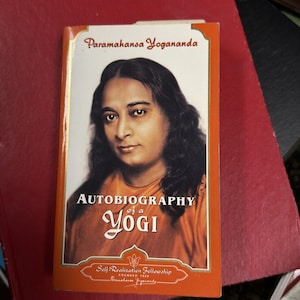 Pode incluir: Um livro intitulado "Autobiografia de um Iogue" de Paramahansa Yogananda. A capa apresenta um retrato do autor em vestes laranja, com o título em uma fonte estilizada. O livro está sobre um fundo vermelho.