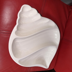 Könnte beinhalten: Weißes Muschelförmiges Serviertablett mit drei Fächern. Das Tablett hat eine glänzende Oberfläche und liegt auf einer roten Oberfläche. Das Tablett ist so gestaltet, dass es einer Muschel ähnelt.