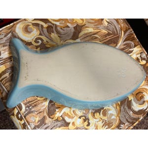 May include: A light blue ceramic fish-shaped serving dish. The dish has a smooth, off-white interior and a light blue exterior with a raised edge. The dish is approximately 12 inches long and 6 inches wide.