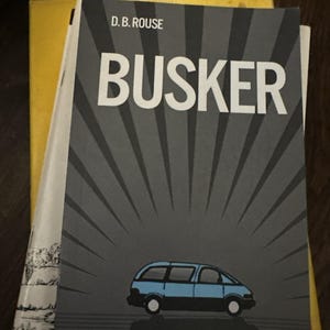 Può includere: Una pila di libri, tra cui un libro intitolato "BUSKER" di D.B. Rouse. La copertina presenta un'illustrazione di un minivan blu su sfondo grigio con linee radianti. Altri libri sono visibili.