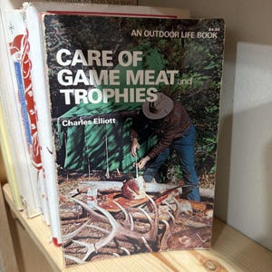 Könnte beinhalten: Ein Vintage-Buch mit dem Titel "Care of Game Meat and Trophies" von Charles Elliott. Das Cover zeigt einen Mann mit Hut, der Fleisch in der Nähe eines grünen Zeltes zubereitet. Das Buch gehört zur Reihe "An Outdoor Life Book".