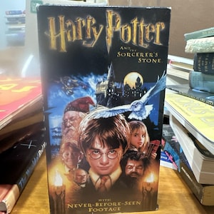 Può includere: Una videocassetta VHS di "Harry Potter e la pietra filosofale" con il titolo in lettere dorate. La copertina presenta un collage di personaggi, il castello di Hogwarts e un gufo. Il testo "NEVER-BEFORE-SEEN FOOTAGE" è in basso.