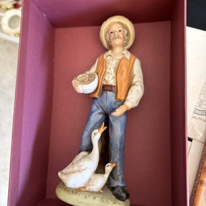 Puede incluir: Figura de cerámica de un anciano con sombrero de paja y chaleco, sosteniendo una cesta, de pie con dos gansos blancos. El hombre viste camisa blanca, vaqueros azules y zapatos negros. La figura está dentro de una caja granate.