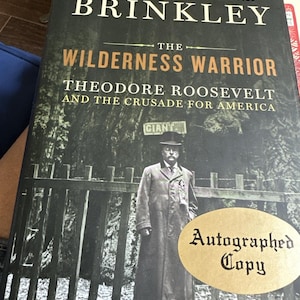 May include: Hardcover book titled "The Wilderness Warrior" by Douglas Brinkley, featuring Theodore Roosevelt. The cover is dark with gold lettering and a sepia-toned image of Roosevelt. An "Autographed Copy" sticker is on the cover.