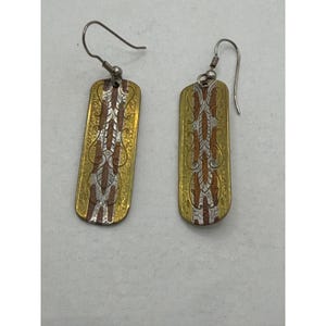 May include: A pair of rectangular drop earrings featuring a hammered gold-tone finish with a central vertical inlay of copper and silver-tone metal. The design includes intricate engraved scrollwork patterns and silver-tone metal ear wires.