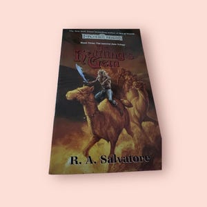 May include: The cover of a fantasy novel titled "The Halflings Gem" by R.A. Salvatore. The cover features a character riding a camel with a sword in hand. The book is part of the Forgotten Realms series and the Icewind Dale trilogy.