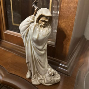 Op de afbeelding: Een keramieken beeldje van een oude man in een gehuurde mantel, met een staf. De figuur is voornamelijk gebroken wit met gouden accenten, en een klein dier rust aan zijn voeten. Het beeldje staat op een houten oppervlak.
