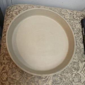 May include: A round, off-white ceramic baking dish with a raised edge. The dish is empty and appears to be unused. The neutral color and simple design suggest it is for baking or serving food. The dish is sitting on a patterned surface.
