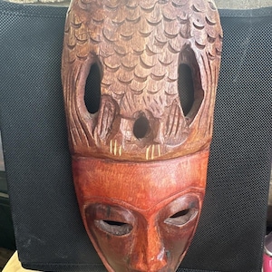 Könnte beinhalten: Eine geschnitzte Holzmaske mit einem Gesicht und eulenartigen Merkmalen. Die Maske ist hauptsächlich braun und rotbraun, mit geschnitzten Details, einschließlich Augen- und Mundöffnungen. Der obere Teil hat ein strukturiertes Muster, das Federn ähnelt.