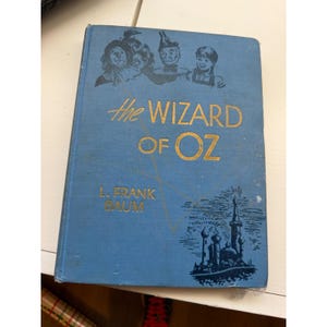 May include: A blue hardcover book titled The Wizard of Oz by L. Frank Baum. The front cover features gold embossed lettering, a black illustration of four characters at the top, and a black graphic of a castle at the bottom right corner.