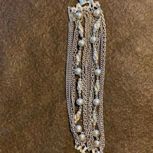 Könnte beinhalten: Silberfarbene Halskette mit mehreren Strängen und verschiedenen Kettentypen. Die Halskette hat kleine, runde Perlen und dekorative Elemente. Die Ketten sind geschichtet und hängen vertikal, wodurch ein strukturiertes und auffälliges Design entsteht.