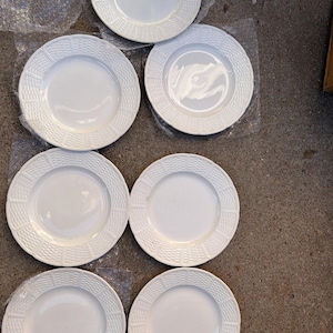 Peut inclure: Ensemble de six assiettes en céramique blanche. Chaque assiette présente un bord texturé, semblable à du tissage, entourant un centre lisse et circulaire. Les assiettes sont disposées sur une surface en plastique transparent.
