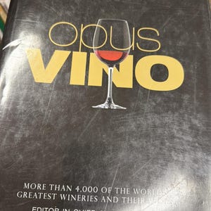 Op de afbeelding: Een boek met de titel "Opus Vino" met een afbeelding van een wijnglas. De zwarte kaft heeft gouden letters. De tekst luidt: "MORE THAN 4,000 OF THE WORLD'S GREATEST WINERIES AND THEIR WINES". Editor-in-Chief is Jim Gordon.