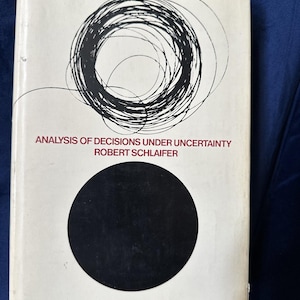 May include: A hardcover book titled "Analysis of Decisions Under Uncertainty" by Robert Schlaifer. The cover features a black circle and a black, scribbled circular design. The book's spine is red.
