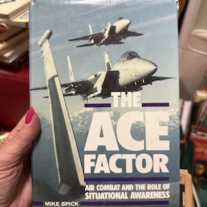 May include: Book cover with the title "The Ace Factor: Air Combat and the Role of Situational Awareness" by Mike Spick. The cover features two fighter jets flying in formation against a blue sky with white clouds.