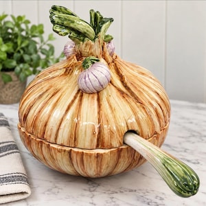 May include: A ceramic tureen shaped like a large onion with a tan and brown textured exterior. The lid features sculpted green onion stalks and small purple-tinted bulbs. A matching ceramic ladle with a green handle extends from the side.