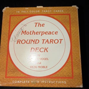 May include: A square, vintage tarot card box with a tan background and a circular design in the center. The design features the words "The Motherpeace ROUND TAROT DECK" and the names of the authors. The box reads "78 FULL-COLOR TAROT CARDS".