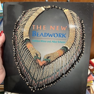 May include: A book titled "The New Beadwork" by Kaitlyn Moss and Alice Scherer. The cover features a close-up of a beaded necklace with a fringe design in shades of gold, black, green, pink, and orange.