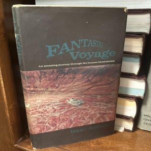 May include: A hardcover book titled "Fantastic Voyage" with a dark brown cover and teal lettering. The cover features an illustration of a spaceship in a red and brown landscape. The author's name, Isaac Asimov, is at the bottom.