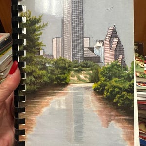 May include: A watercolor painting of a city skyline with a river in the foreground. The buildings are tall and modern, with a few trees in the foreground. The sky is a light blue with a few clouds. The painting is signed "Tina Goldsmith © 93".