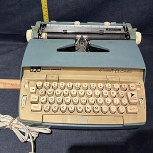 May include: A vintage Smith-Corona Coronet Automatic typewriter in a light blue and beige color scheme. The keyboard features round, off-white keys with black lettering. The typewriter has a power return key and a cord.