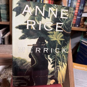 May include: A hardcover book titled "Merrick" by Anne Rice. The cover features a dark silhouette figure playing a flute, set against a backdrop of lush green foliage. The title and author's name are in white, with the word "Merrick" prominently displayed.
