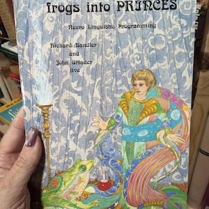 Puede incluir: Portada del libro "frogs into PRINCES" con el subtítulo "Neuro Linguistic Programming". La ilustración muestra una rana, una mujer con un vestido y una botella de poción. Autores: Richard Bandler y John Grinder.