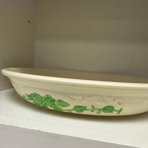 Może przedstawiać: Owalna ceramiczna miska do serwowania w kolorze ecru z zielonym kwiatowym wzorem. Miska ma lekko podniesiony brzeg i jest ozdobiona powtarzającym się wzorem kwiatów i liści. Miska stoi na białej półce.