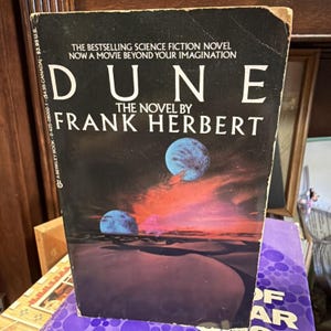 May include: A vintage paperback book titled "Dune" by Frank Herbert. The cover features a dark background with a desert landscape and two blue planets. The title is in large white letters, with additional text at the top.