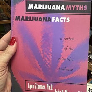 Può includere: Un libro intitolato "Marijuana Myths Marijuana Facts" con una copertina rosa e viola. La copertina presenta testo e un design stilizzato a forma di foglia di cannabis. Il libro è una revisione delle prove scientifiche.