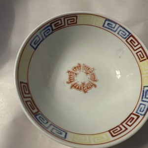 Peut inclure: Un bol en céramique blanche avec un motif géométrique rouge, jaune et bleu autour du bord. Le bol a un motif floral rouge et blanc au centre.