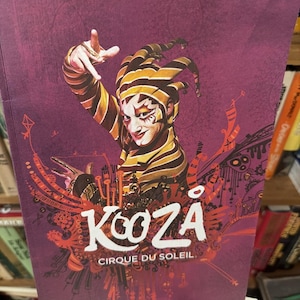 Puede incluir: Una portada de programa púrpura para el espectáculo Kooza del Cirque du Soleil. La portada presenta a un artista con un disfraz de arlequín con un sombrero a rayas amarillas y negras y una cara pintada. El título del espectáculo está en letras blancas grandes.