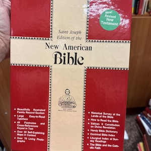 May include: A red and cream-colored book titled "New American Bible" with the text "Saint Joseph Edition of the" above it. The cover includes a "New! Revised New Testament" sticker. Features include footnotes, cross-references, and a family record section.
