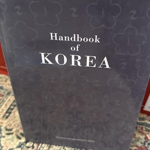 Könnte beinhalten: Ein dunkelblaues Hardcover-Buch mit dem Titel "Handbook of KOREA" und weißem Text. Das Cover zeigt ein dezentes Blumenmuster. Das Buch scheint neu und in gutem Zustand zu sein.