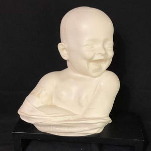 May include: A cream-colored bust of a laughing baby. The sculpture depicts a bald baby with a wide, open-mouthed smile. The baby's shoulders and chest are visible, with a draped garment partially covering the body. The bust is placed on a black surface.