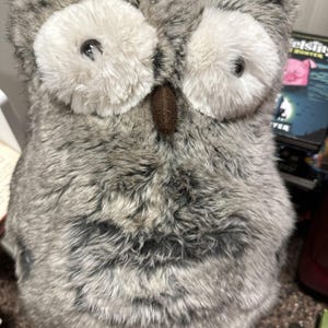 May include: A plush owl toy with gray and white fur. The owl has large white eyes with black pupils and a brown beak. The toy is made of soft, textured material and is designed to resemble a real owl.