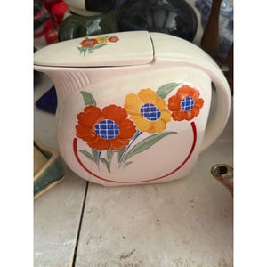 May include: A cream-colored ceramic pitcher with a matching lid, featuring a hand-painted floral design of two large orange flowers and one yellow flower with blue grid centers, accented by green leaves and a thin red circular line border.