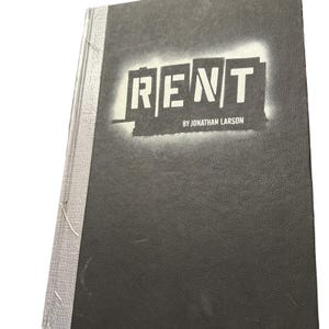 Puede incluir: Un libro de tapa dura negro con el título "RENT" en letras blancas estilizadas. El nombre del autor, "BY JONATHAN LARSON", está impreso debajo. El lomo del libro es de color gris claro.