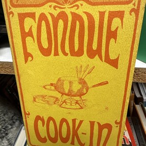 Può includere: Un libro di cucina vintage con sfondo giallo e testo arancione che recita "FONDUE COOK-IN". La copertina presenta un'illustrazione di una fonduta con forchette. Il libro è una guida alla cucina della fonduta.