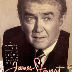Puede incluir: Portada de libro con un retrato de James Stewart. El título es "A Wonderful Life: The Films and Career of James Stewart" en escritura blanca y rosa. El libro es una biografía del actor.