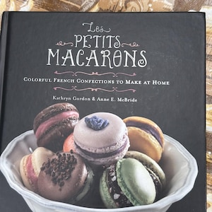Peut inclure: Un livre de cuisine intitulé "Les Petits Macarons" avec des macarons colorés dans un bol blanc. La couverture du livre est noire avec du texte blanc, et les macarons sont de couleurs variées, dont rose, marron et vert. Le sous-titre du livre est "Colorful French Confections to Make at Home."