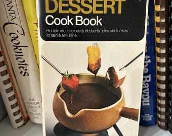May include: A vintage cookbook titled "Pillsbury's DESSERT Cook Book" with a black and gold cover. The cover illustration shows a fondue pot with dipping fruit. Other cookbooks are visible in the background.