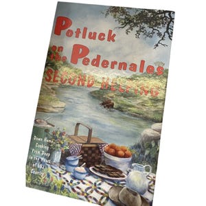May include: A vintage cookbook cover titled "Potluck on the Pedernales, Second Helping." The cover features a picnic scene with a river, food, and text. The text includes "Down Home Cooking From Deep in the Heart of LBJ Country."