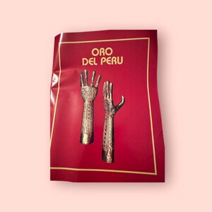 Op de afbeelding: Een rode kaart met gouden rand en de tekst "ORO DEL PERU" in gouden letters. De kaart toont twee gedetailleerde, metaalachtig uitziende handsculpturen.