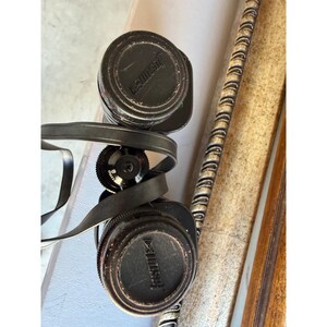 May include: Black binoculars featuring circular lens caps with the brand name Tasco embossed on the surface. The device includes a central focus adjustment wheel and a black neck strap wrapped around the body of the optical instrument.