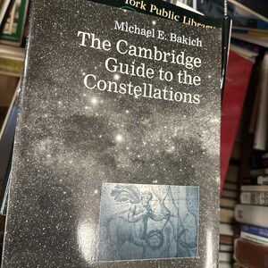 Puede incluir: Un libro titulado "The Cambridge Guide to the Constellations" de Michael E. Bakich. La portada presenta un diseño de cielo estrellado con texto blanco. Una ilustración azul de una figura mitológica está en la parte inferior derecha.